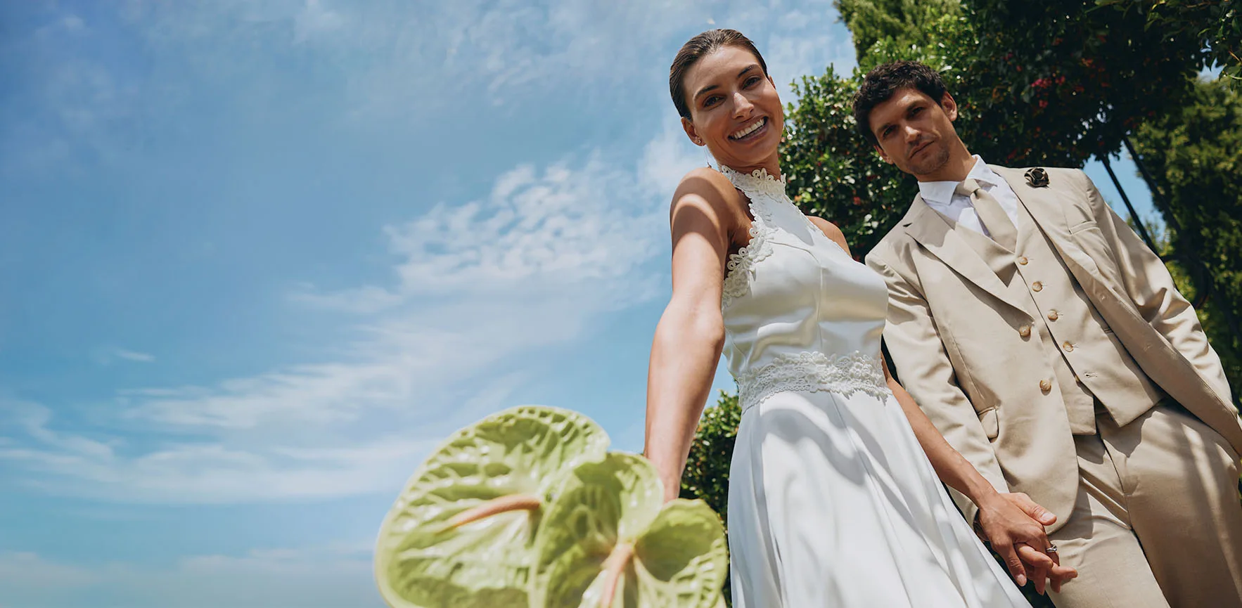 Elegantný svadobný pár na slnku: ona v bielych šatách, on v béžovom obleku so slnečnými okuliarmi.